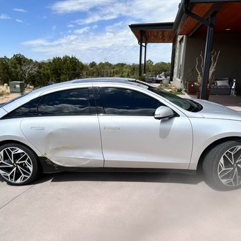 Body Master - A silver sedan with tinted windows and stylish alloy wheels is parked on a driveway in front of a house. The vehicle has visible damage, including dents and scratches on the driver's side door and rear panel. The background features a house with a porch, outdoor furniture, and a landscaped yard with desert plants and trees. The sky is partly cloudy with patches of blue. - Body Shops Near Me - Albuquerque, New Mexico