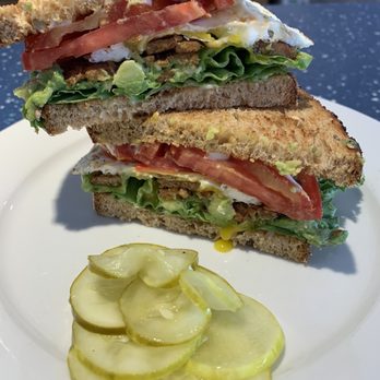 Common Roots - TLT w avocado and egg. - A sandwich featuring two slices of toasted bread filled with lettuce, tomato slices, a fried egg, avocado, and a protein that appears to be beef or sausage. The sandwich is cut in half and stacked, served on a white plate with sliced pickles on the side. The background suggests a casual dining setting. - Cafes Near Me - Rye, New Hampshire