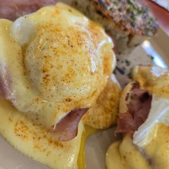 Snooze, an A.M. Eatery - Eggs Benedict served with hollandaise sauce, ham, and seasoning, accompanied by a piece of toast with herbs. - Breakfast & Brunch Near Me - Gilbert, Arizona