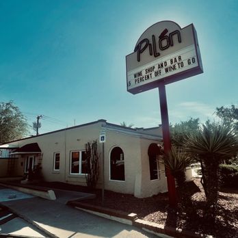 Pilon - a view of the front of a restaurant - The image shows the exterior of Pilon, a wine shop and bar, with a large vertical sign displaying the business name. The building is a single-story structure with a light-colored facade and arched windows. Surrounding the building are desert plants, including yucca and other drought-tolerant shrubs. A parking lot with a white pickup truck is visible in the foreground, and the sky is clear with a few clouds, indicating a sunny day. The scene is set in a commercial area with other buildings and traffic in the background. - Beer, Wine & Spirits Near Me - El Paso, Texas