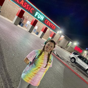 Classic Fun Center - A smiling young woman with braided hair, wearing a colorful tie-dye shirt and patterned pants, stands in front of Classic Fun Center at night. - Party & Event Planning Near Me - Sandy, Utah