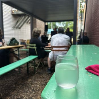 Table Wine - Patio - A clear glass of water is positioned on a green painted outdoor table, with a pink cloth or napkin nearby. In the background, several people are seated at tables, engaged in a gathering or event, under a shaded structure with a corrugated roof. The setting appears to be an outdoor patio or garden area, with some trees and buildings visible beyond the gathering. - Beer, Wine & Spirits Near Me - Madison, Wisconsin