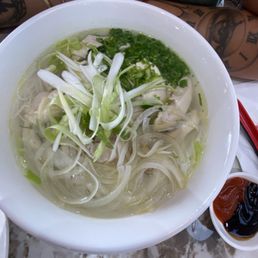 Chicken Pho - A large white bowl contains chicken pho, a Vietnamese noodle soup with clear broth, rice noodles, sliced chicken, and garnished with chopped green onions and shredded cabbage. The bowl is placed on a marble table, with side sauces visible nearby. The image includes part of a person's arm and tattoos in the background.