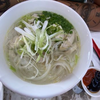 Lotus Tea - Chicken Pho - A large white bowl contains chicken pho, a Vietnamese noodle soup with clear broth, rice noodles, sliced chicken, and garnished with chopped green onions and shredded cabbage. The bowl is placed on a marble table, with side sauces visible nearby. The image includes part of a person's arm and tattoos in the background. - Bubble Tea Near Me - Scranton, Pennsylvania