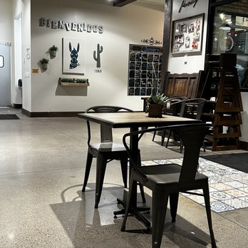 Someburros - A wooden table with two black chairs is positioned in a restaurant or cafe interior. The table has a small potted plant as a centerpiece. The background features a wall with decorative elements, including a sign that says 'BIENVENIDOS', framed photos, and a chalkboard menu. There are additional chairs stacked against the wall, and the floor is a polished concrete surface. The lighting is soft, creating a cozy atmosphere. - Mexican Near Me - Mesa, Arizona