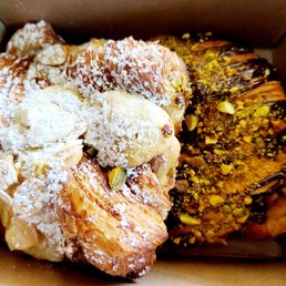 Pistachio Croissant - A variety of pastries are shown inside a cardboard box, including one dusted with powdered sugar and topped with chopped nuts, and another covered with chocolate and sprinkled with chopped pistachios. The pastries appear freshly baked and are arranged closely together.