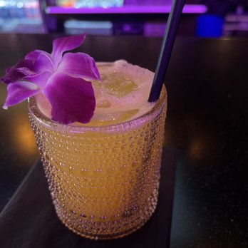Killer Queen - Charlemagne drink - A yellow cocktail with ice cubes is presented in a textured glass, garnished with a purple flower and a black straw. The drink is placed on a dark bar table with a black coaster underneath. The background features a bar setting with purple lighting and blurred shelves. - Pizza Near Me - Cincinnati, Ohio