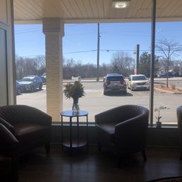Tea and dessert seating area. - A cozy indoor seating area features four leather chairs, a small round table with a flower vase, and a large window overlooking a parking lot on a sunny day.