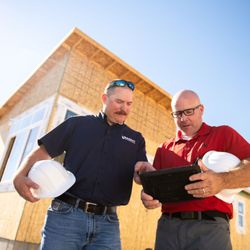 Builders FirstSource - Two Builders FirstSource team members hold hard hats while reviewing construction plans on a tablet...