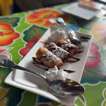 Kai Yang - Fried Banana - Four rolled desserts are served on a rectangular white plate, garnished with powdered sugar and chocolate drizzle, with a dollop of whipped cream. - Thai Near Me - Montclair, New Jersey