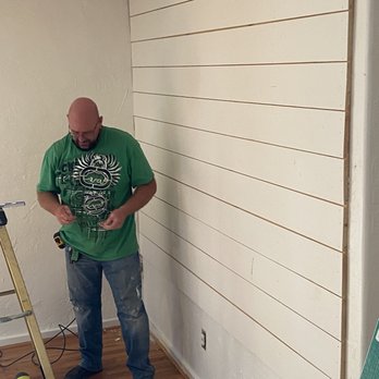 Knotty's Custom Carpentry - Working his magic. - A man with a bald head and glasses is standing in a room, working on a wall that has horizontal wooden slats attached to it. He is wearing a green T-shirt, jeans, and work shoes, and appears to be holding tools or hardware. The room has hardwood flooring, a ladder, and some tools on the floor, indicating ongoing construction or renovation work. An electrical outlet is visible on the wall below the slats. - Handyman Near Me