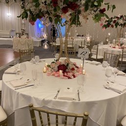 Prof Party Decor - A round banquet table features a tall floral centerpiece with pink, white, and red flowers, surrounded by candles and table settings in an event hall.