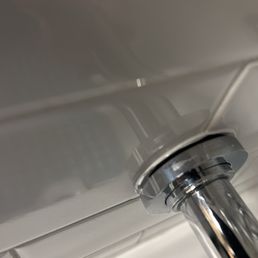 A close-up image of a metallic toilet flush handle mounted on a toilet tank. The background shows white tiled walls, indicating a bathroom setting. The handle is shiny and reflective, with the tank surface smooth and clean.