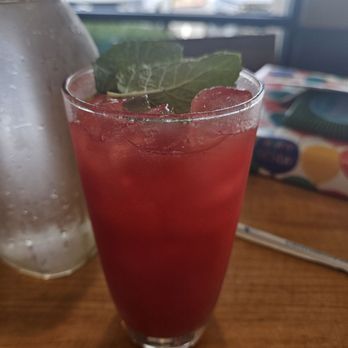 First Watch - Watermelon refresher (seasonal menu June 2024) - A tall glass filled with a red watermelon refresher, ice cubes, and garnished with a mint leaf, on a wooden table. - Breakfast & Brunch Near Me - Gilbert, Arizona