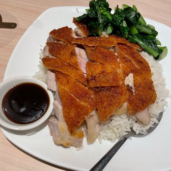Congee Queen - Roast pork belly rice - The image shows a white plate containing slices of crispy roast pork belly placed on a bed of white rice. There are sautéed green vegetables, likely bok choy or similar greens, on the side. A small bowl of soy sauce is positioned on the plate for dipping. The plate is set on a light-colored wooden table, and a metal spoon rests on the edge of the plate. - Chinese Near Me - Markham, Ontario