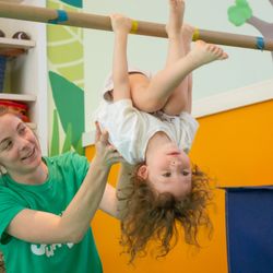 Jump! Gymnastics - A coach supports a young gymnast as he hangs upside down on the bar.