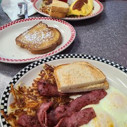 Ze's breakfast - The image shows a breakfast setting with a plate containing fried eggs, strips of bacon, toasted bread, and hash browns. In the background, there is another plate with an omelet topped with sauce, slices of bread, and a small container of jam. The table has a textured surface, and a person is partially visible, holding a fork and knife. The dishes are served on plates with checkered borders, typical of a diner setting.