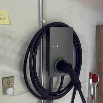 Jon Dyer Electrician - nice clean EV charger installed very close to my car's charging port & garage door in case i need to stretch the cable outside. - A gray and black electric vehicle charger with a green accent ring is mounted on a wall in a garage or utility area. The charger has a coiled cable hanging from it, with a connector attached. Nearby, there is an electrical outlet, a conduit running vertically, and a small white box mounted on the wall. The wall shows some signs of wear and minor damage, and there are miscellaneous items such as a wooden board and a red reflector in the background. - Electricians Near Me