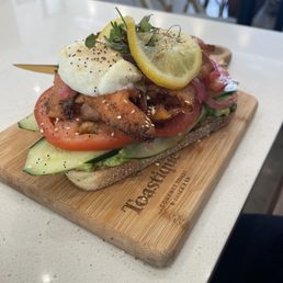Salmon Pastrami Toast - A sandwich on a wooden serving board contains sliced tomato, cucumber, grilled meat, and a dollop of cream or cheese, topped with lemon slices and microgreens. The sandwich is assembled on a piece of bread, with visible seasoning and garnishes, and is presented on a branded wooden board in a casual dining setting.