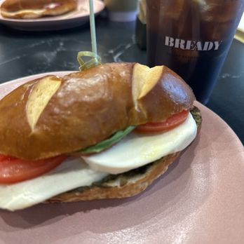 Bready Cafe - Pretzel bun caprese sandwich - A sandwich featuring a pretzel bun, fresh tomato, basil, and mozzarella cheese, served on a pink plate with a drink in the background. - Bakeries Near Me - Toronto, Ontario