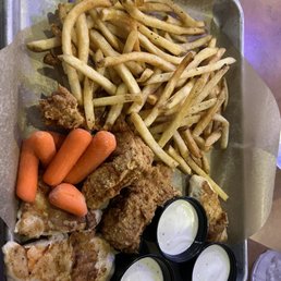 Chicken Tenders - A metal tray lined with parchment paper holds fried chicken pieces, a generous serving of French fries, small cooked baby carrots, and three small cups of white dipping sauce. The tray is placed on a table with a menu or paper partially visible in the background. The food appears to be typical of American casual dining or bar food.