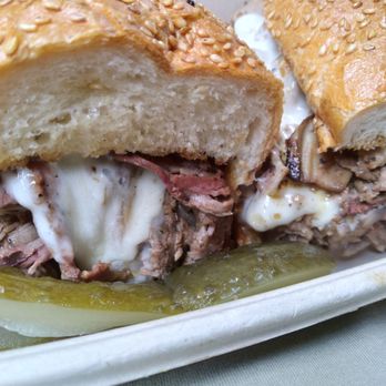 Stack'd Deli Kitchen - A sandwich featuring sliced roast beef and melted cheese inside a sesame seed bun is shown in a takeout container. The sandwich is cut in half, revealing the ingredients, and is accompanied by pickles and onions at the base. The bread has a golden-brown crust with sesame seeds on top. - Salad Near Me - Toronto, Ontario