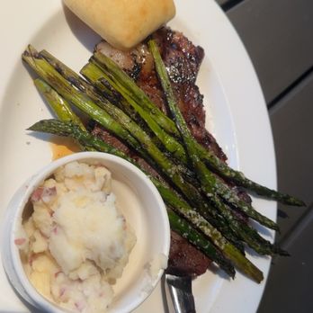 Harveys - Marinated Ribeye (Dinner 4pm-Close) - A dinner plate contains grilled asparagus spears, a baked bread roll, mashed potatoes with bits of bacon or ham, and a cooked steak. The food is arranged on a white oval plate, with the steak partially covered by the asparagus and the mashed potatoes in a small cup. The presentation suggests a hearty meal served in a casual dining setting. - Seafood Near Me - Columbus, Mississippi