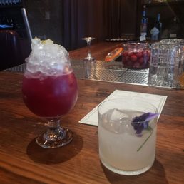 Inner Clarity - The image shows two cocktails on a wooden bar counter in a dimly lit bar. The first cocktail is in a tall glass with crushed ice, a straw, and a lemon wedge. The second cocktail is in a shorter glass, garnished with a purple flower and ice cubes. The background includes bottles of alcohol, glasses, and bar equipment, with a dark curtain and shelves behind the bar.