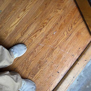 Arch Moving - Scratch in hardwood - The image shows a person's feet in white sneakers and beige pants standing on a wooden floor with visible scratches and small debris. The floor transitions to a concrete surface near a threshold, with part of a black appliance or furniture visible in the upper right corner. The photo captures the lower legs and feet, focusing on the condition of the wooden flooring. - Movers Near Me - Saint Ann, Missouri