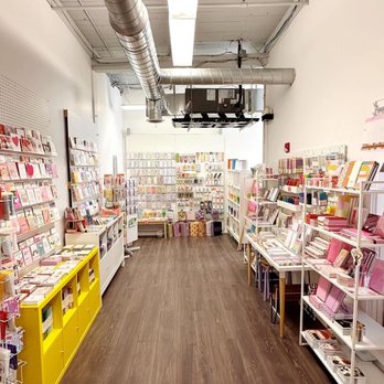 Calliope Paperie - Stationery Fans are happy here! - The image shows the interior of a stationery and gift shop with white walls and a wooden floor. Shelves on both sides are stocked with greeting cards, notebooks, washi tape, and various small gift items. The ceiling has exposed ductwork and a large fluorescent light fixture. The shop appears well-organized and brightly lit, with a spacious aisle in the center for browsing. - Gift Shops Near Me - Hudson, Massachusetts