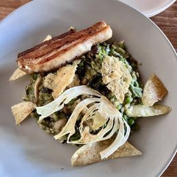 Pork Belly - A plate of mushroom risotto topped with slices of pork belly, accompanied by cauliflower florets and crispy chips. The dish is presented on a white, round plate, with a wooden table surface visible in the background. The risotto appears creamy with visible mushroom pieces, and the pork belly has a crispy exterior.