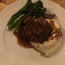 Filet Mignon - A dinner plate displays a filet mignon topped with gravy, served alongside mashed potatoes with gravy, steamed broccoli, and sautéed mushrooms. The dish is presented on a white, decorative plate on a wooden table.