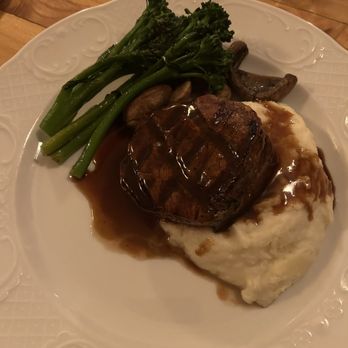 The Orville - Filet mignon - A dinner plate displays a filet mignon topped with gravy, served alongside mashed potatoes with gravy, steamed broccoli, and sautéed mushrooms. The dish is presented on a white, decorative plate on a wooden table. - Cocktail Bars Near Me - West Bend, Wisconsin