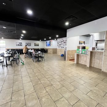 Tacarbon - Other dining area with TV - The image shows a large, mostly empty dining area with tiled flooring and a black ceiling with recessed lighting. There are several black chairs and tables, some occupied by a person and a child. A TV is mounted on the white wall, displaying content. The space includes a counter area with drink dispensers and a window for food service. The overall setting appears to be a casual restaurant or cafe. - Mexican Near Me - Peoria, Arizona