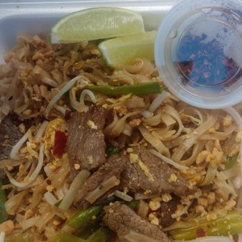 Thai Tip - Pad Thai noodles beef - A takeout container holds Pad Thai noodles with slices of beef, bean sprouts, chopped green onions, and crushed peanuts. Two lime wedges are placed on top for garnish, and a small clear cup contains a dark sauce, likely for dipping or flavoring. The dish appears freshly prepared and ready for consumption, typical of Thai cuisine. - Thai Near Me - Albuquerque, New Mexico