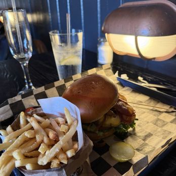 J Murphys on Main - French fries, bacon cheeseburger with grilled onions - A burger with a toasted bun, lettuce, bacon, and other toppings is placed on a checkered paper-lined tray. Accompanying the burger are French fries in a paper container, with sliced pickles on the tray. The setting appears to be a restaurant with a dark wooden table, a blue wall, and a warm light fixture overhead. Two drinks, one with a straw and lemon, are visible in the background. - American Near Me - Cold Spring, New York