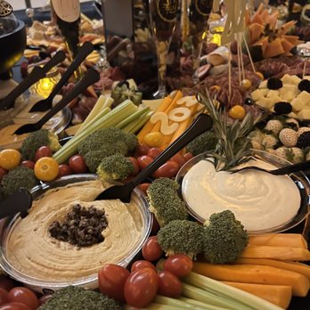 M Events - A food display includes fresh vegetables such as cherry tomatoes, celery sticks, broccoli, and cheese slices, along with dips and spreads. There are also charcuterie items, cheese boards, and decorative elements like balloons and flowers. The setup appears to be for a celebratory event or gathering, with a variety of food options and drink glasses visible in the background. - Caterers Near Me