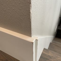 The image depicts a close-up of a corner of a white baseboard trim with chipped paint and minor damage. The baseboard is positioned against a textured wall painted in a beige color, with a hardwood floor visible at the bottom. The focus is on the area where the baseboard meets the wall, highlighting imperfections and wear.