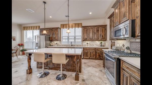 Kitchen Cabinet Remodel - This kitchen remodel features rich knotty alder wood cabinets in a dark stain, showcasing natural knots and grain for a rustic charm. The spacious layout includes upper and lower cabinets framing a window with valance, a tiled backsplash in neutral earth tones, and stainless steel appliances like a side-by-side refrigerator, over-the-range microwave, and gas stove. A central island with white quartz countertop seats two beige swivel stools under pendant lights, all set on large travertine-style floor tiles for a warm, inviting space.