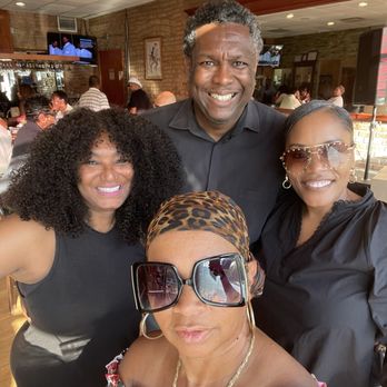 Norman's Bistro - Group photo - A group of four people, three women and one man, are taking a selfie inside a restaurant with brick walls and a wooden trim. The woman in the foreground wears large sunglasses, a colorful off-shoulder top, and a headscarf. The woman on the left has curly hair and is smiling, while the woman on the right wears glasses and a black top. The man in the back is smiling and dressed in a black shirt. The restaurant has a lively atmosphere with other diners and a television screen visible in the background. - Brazilian Near Me - Chicago, Illinois