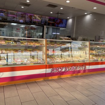 Amy's Donuts - Look at the selection! - A glass display case showcases a variety of donuts and baked goods at Amy's Donuts in Denton, Texas. The shop features a colorful exterior with the business name prominently displayed. Digital menu boards are visible above the counter, listing donut options, beverages, and prices. The interior includes tiled flooring and a large window allowing natural light to enter, with a view of the outside street scene. - Donuts Near Me - Denton, Texas