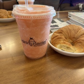 Raining Berries - Passion Berry Smoothie - A Passion Berry smoothie in a clear plastic cup with a straw, placed on a wooden table at a café, with a croissant on a white plate nearby. - Acai Bowls Near Me - Brandon, Florida