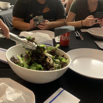 Selfup - Mozzarella, Balsamic dressing and arugula - Several individuals are seated at a dining table, each engaged with their smartphones. The table has a large bowl of salad, a Coca-Cola can, and various plates and utensils. The setting appears to be a casual restaurant or dining area, with other diners visible in the background. The image captures a moment of people multitasking during a meal. - Cooking Classes Near Me - Boston, Massachusetts