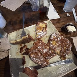 Yum yum - A square plate holds several pieces of French toast dusted with powdered sugar, with some bacon pieces nearby, on a wooden table.