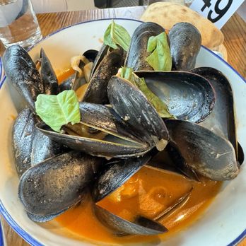 Boschetto - A white bowl with a blue rim contains cooked mussels in a red tomato sauce, garnished with fresh basil leaves. The dish is served on a wooden table, with a glass of water, a piece of bread, and a table number card visible in the background. The setting appears to be a casual dining restaurant. - Italian Near Me - Montclair, New Jersey