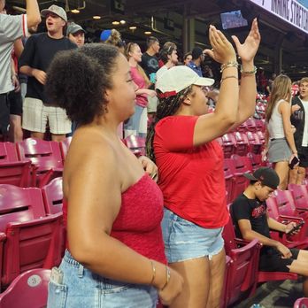Cincinnati Reds - Two women are standing in stadium seats, actively cheering during a baseball game. One woman is wearing a red strapless top and denim shorts, while the other is in a red t-shirt, denim shorts, and a white cap. They are surrounded by other spectators, some standing and some seated, with a large digital scoreboard and advertisements visible in the background. The scene captures a lively moment at a baseball game. - Professional Sports Teams Near Me - Cincinnati, Ohio