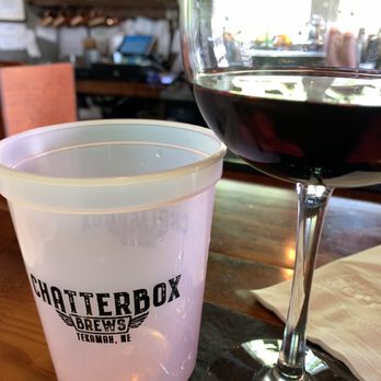 Chatterbox Brews - a glass of wine and a glass of wine - A glass of red wine and a plastic cup with the Chatterbox Brews logo are placed on a wooden table inside a restaurant or bar. - Venues & Event Spaces Near Me - Tekamah, Nebraska