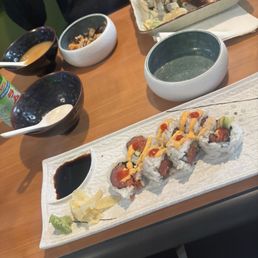 Spicy Tuna Roll - Sushi rolls topped with sauces and accompanied by soy sauce, wasabi, and pickled ginger on a rectangular plate at a dining table.