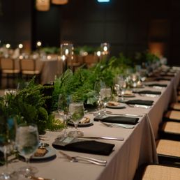 A long rectangular dining table is arranged for a formal event, decorated with abundant green foliage and plants as a centerpiece. The table is set with wine glasses, water glasses, plates, and black cloth napkins, with silverware placed neatly. The room features warm lighting, with additional tables visible in the background, and a dark, elegant ambiance suitable for a celebration or gathering.