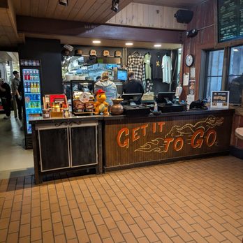 Dinosaur Bar-B-Que - A restaurant or cafe counter with a sign that says "GET IT TO GO" in orange letters. Behind the counter, staff are working, with a menu screen visible on the right. Merchandise, including shirts and hats, are displayed on the wall. The area has a wooden ceiling and brick flooring, with a refrigerator stocked with drinks on the left side. - Barbeque Near Me - Troy, New York